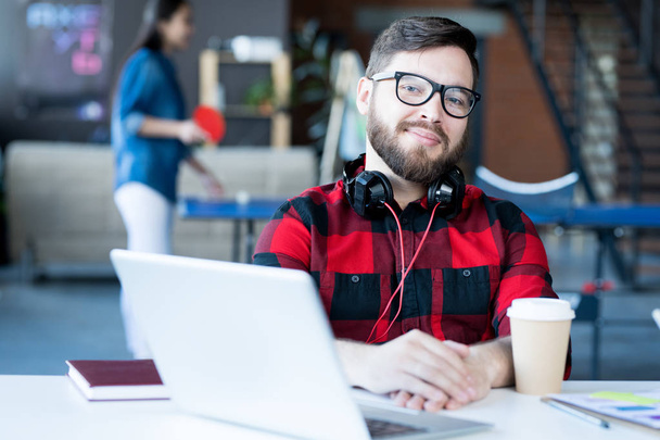 Portrét usmívajícího se vousatého muže ve sluchátkách a brýlích, které se dívají na kameru, zatímco sedí u stolu v moderní kanceláři developerského podniku, kopírovat prostor - Fotografie, Obrázek