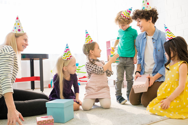 Cheerful young mothers in casual clothing sitting on carpet and playing with kids at birthday party, they looking after children while children giving gifts to birthday girl - 写真・画像
