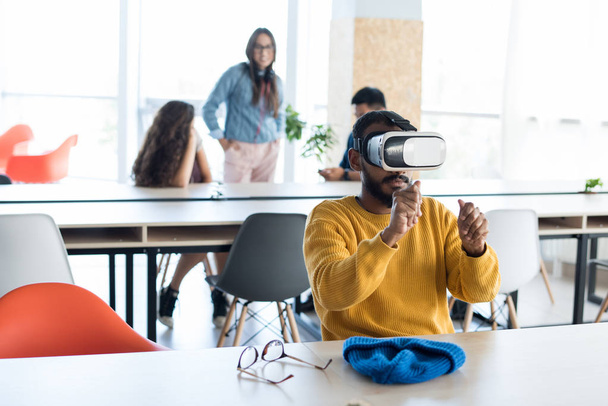 Serious curious young middle-eastern hipster man with beard sitting at table and holding invisible object while playing video game in VR headset - Photo, Image