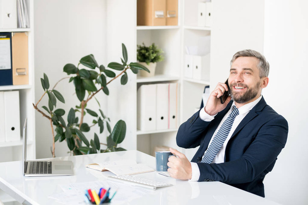 Cheerful excited handsome bearded businessman sitting at table with diary and computer in modern office and communicating on mobile phone while drinking coffee - Fotoğraf, Görsel
