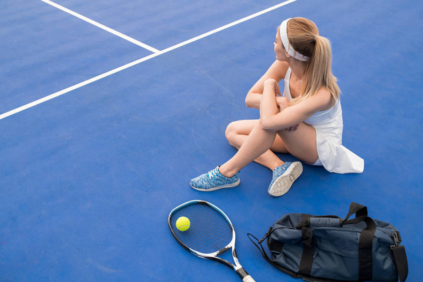 Wysoki kąt Portret młodej Blondynki kobieta siedzi na podłodze w krytym korcie tenisowym, biorąc przerwę od praktyki, Kopiuj przestrzeń - Zdjęcie, obraz