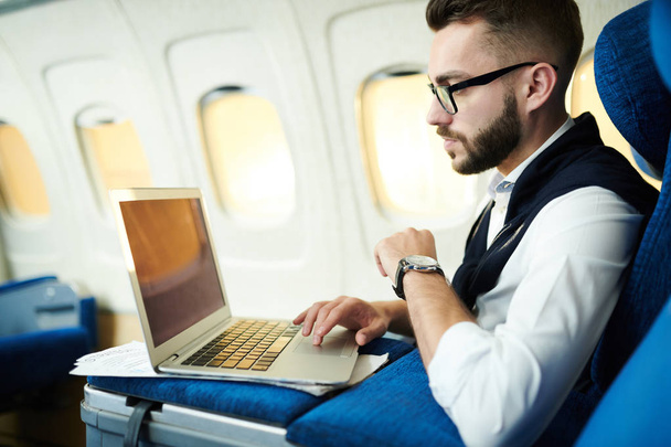Retrato de vista lateral del hombre de negocios guapo que usa el ordenador portátil mientras trabaja en avión durante el vuelo largo de primera clase, espacio de copia
 - Foto, Imagen