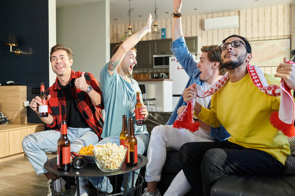 Cheerful young guys are watching match on couch at home and drink beer, they are fans of sports games as football, basketball, hockey, baseball - Photo, Image