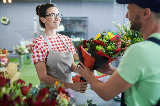Прочекайте портрет усміхненої молодої жінки, яка дарує букет, щоб доставити хлопця в маленький квітковий магазин, копіювати простір
 - Фото, зображення
