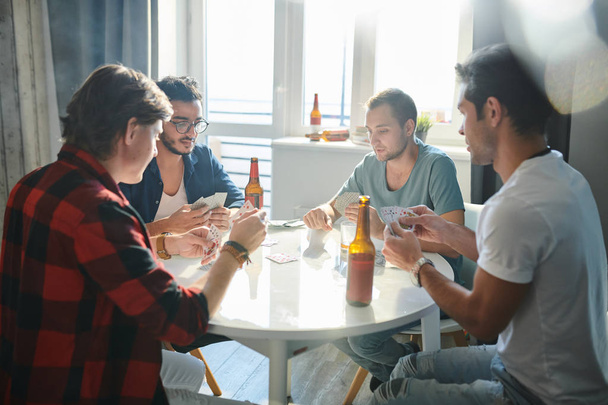 Group of young male friends resting at the table with beer and cards at home party - Photo, Image