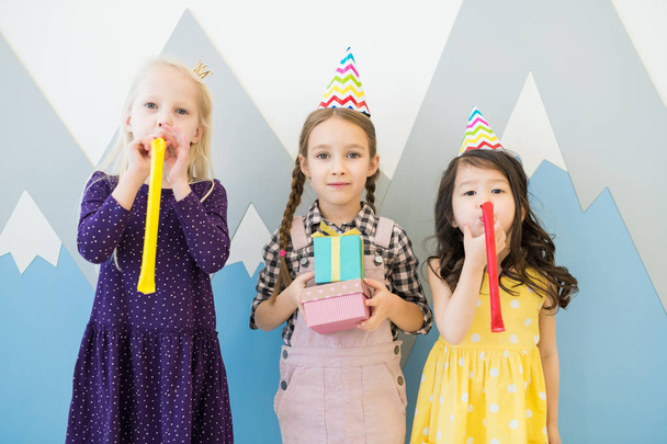 Group of content pretty multi-ethnic girls in casual clothing standing against wall with drawing of mountains and blowing out party horns while having fun at childrens birthday party. - Photo, Image