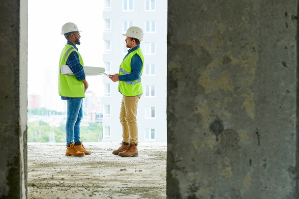 Obsah mladí víceetnické stavební inženýři, kteří nosí bezpečnostní vesty a pracovní přilby stojící na staveništi a pracují na projektu výstavby bytu - Fotografie, Obrázek