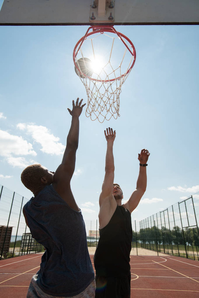Action-Porträt zweier junger Männer, die Basketball spielen und mit einem Reifen gegen den blauen Himmel springen, der von Sonnenlicht erhellt wird - Foto, Bild