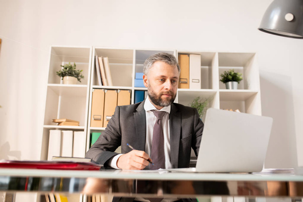Серьезный занятой менеджер в формальном костюме сидит за столом и использует ноутбук при анализе информации в современном офисе
 - Фото, изображение