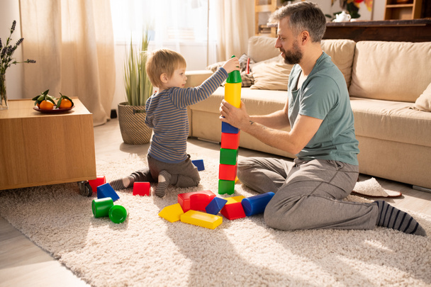 Ciekawy cute little Son w homewear siedzi na dywanie i oddanie stożka na wieży wykonane z kolorowych kostek, brodaty ojciec i syn gry z zabawkami kostki - Zdjęcie, obraz