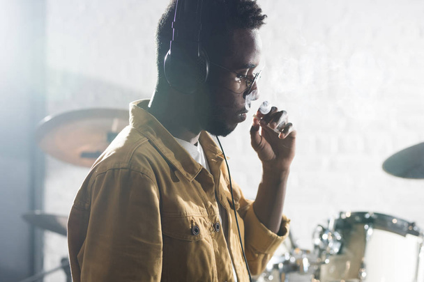 Vista lateral retrato do homem afro-americano contemporâneo fumando durante o ensaio da banda em estúdio de gravação, espaço de cópia
 - Foto, Imagem