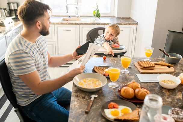 Vista lateral retrato do jovem pai lendo jornal no café da manhã enquanto olha para o bebê bonito sentado em cadeira de crianças por mesa, espaço de cópia
 - Foto, Imagem