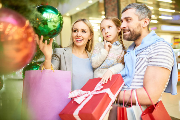 Casal com a filhinha olhando para bugigangas na árvore de Natal enquanto fazem compras juntos
 - Foto, Imagem