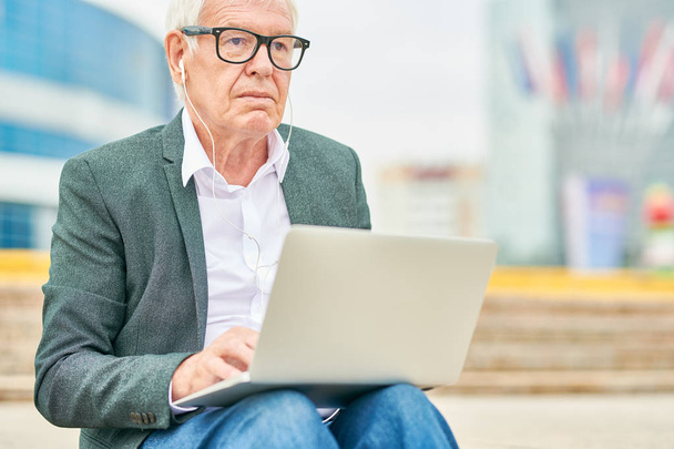 眼鏡をかけた老人がイヤホンで音楽を聴き、通りのぼやけた背景に座りながらノートパソコンを閲覧する - 写真・画像