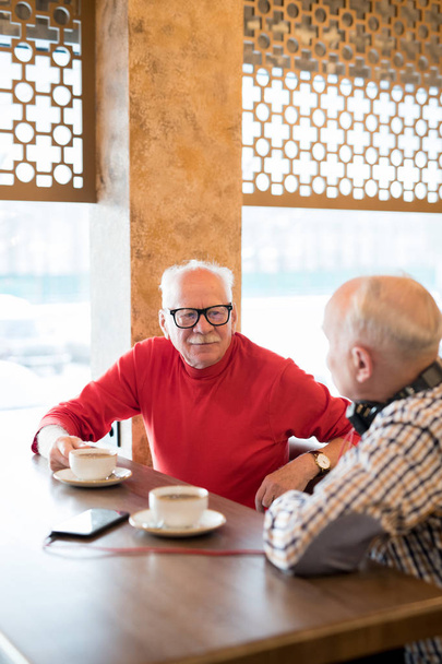 Содержание любопытный красивый пожилой мужчина в красной водолазке и очки, сидя за деревянным столом и разговаривая с другом в кафе
 - Фото, изображение