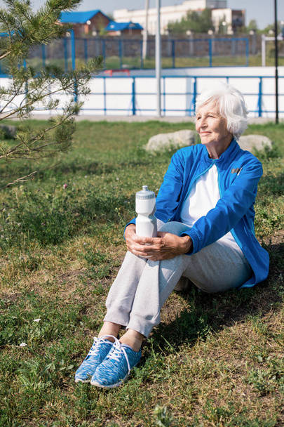 Портрет сучасної старшої жінки, яка сидить на газоні і щасливо посміхається після ранкового бігу в парку
 - Фото, зображення