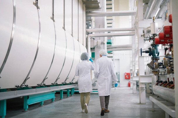 Visão traseira retrato de comprimento total de dois trabalhadores de fábrica vestindo jalecos cruzando oficina de produção, espaço de cópia
 - Foto, Imagem