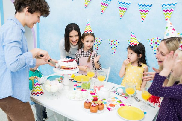 Χαμογελαστή θετική ελκυστική μητέρα σε casual πουκάμισο στέκεται στο τραπέζι με επιδόρπια και κοπή τούρτα με μαχαίρι, ενώ το περιεχόμενο παιδιά σε καπέλα κόμμα τρώγοντας γλυκά - Φωτογραφία, εικόνα