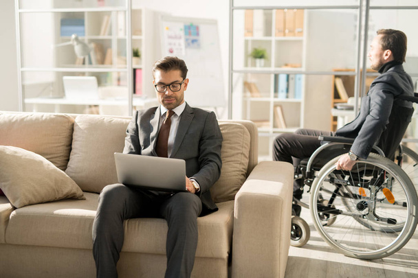 Gerente serio en traje sentado en el sofá y mecanografía en el ordenador portátil con hombre de negocios discapacitado moviéndose por la oficina en silla de ruedas en el fondo
 - Foto, Imagen