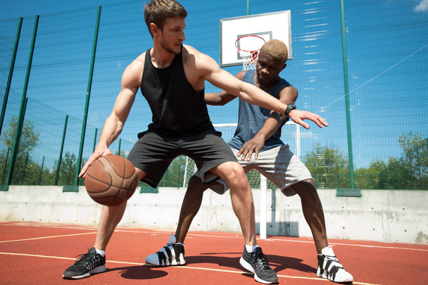 Täyspitkä toiminta laukaus kaksi lihaksikasta nuorta miestä pelaa koripalloa ulkona tuomioistuimessa
 - Valokuva, kuva