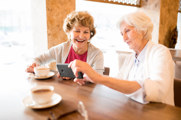 コーヒーカップを持ってテーブルに座り、スマートフォンでビデオを見ながら笑う陽気な興奮した老婦人 - 写真・画像