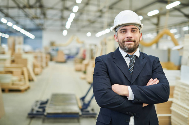 Content confident handsome middle-aged bearded businessman in hardhat crossing arms on chest and looking at camera while standing at furniture factory - Φωτογραφία, εικόνα