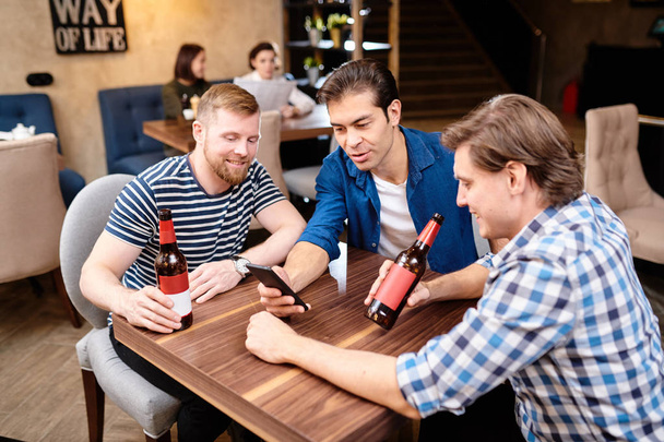 Jolly przystojny męski przyjaciele w casual Odzież siedzący przy stół i przy pomocy smartphone chwila surfing Social Media razem w Bar - Zdjęcie, obraz
