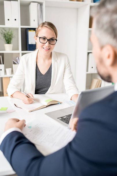 Uśmiechnięta atrakcyjna kobieta konsultant biznesowy w okularach rozmawia z przedsiębiorcą i wyjaśnia podejście do niego, podczas gdy siedzą przy stole w nowoczesnym biurze - Zdjęcie, obraz
