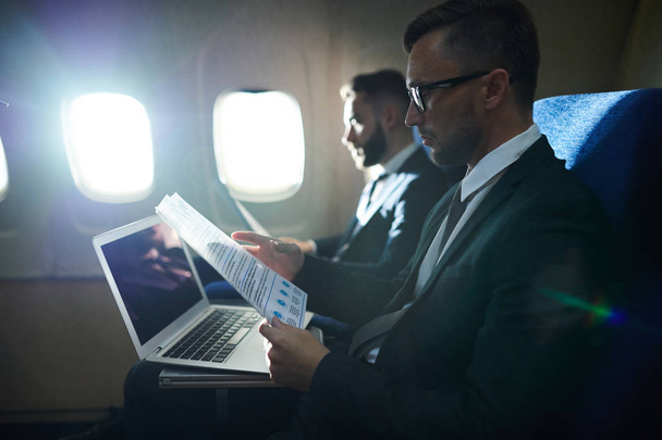 Birinci sınıf uçuş sırasında loş ışıklı düzlemde çalışan ve belgeleri okuyan iki iş adamının yan görünüm portresi, kopya alanı - Fotoğraf, Görsel