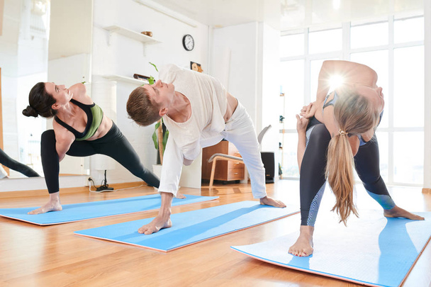 Grupo de jóvenes flexibles parados en colchonetas de ejercicio en fila y tomados de la mano a sus espaldas y torciendo la parte superior del cuerpo a un lado en la clase de yoga
 - Foto, Imagen