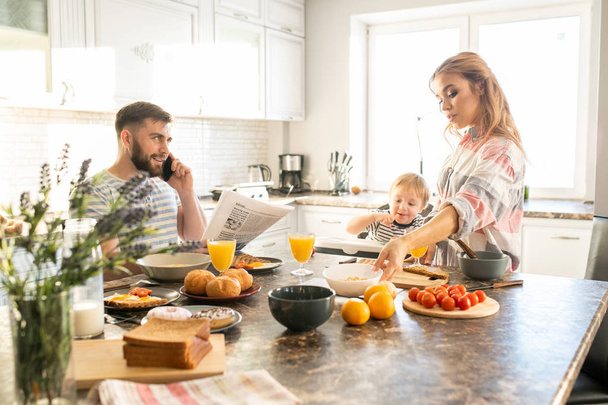 Portrait of happy family enjoying breakfast in kitchen, young parents with cute baby sitting in kid chair at big table, copy space - Foto, Imagen