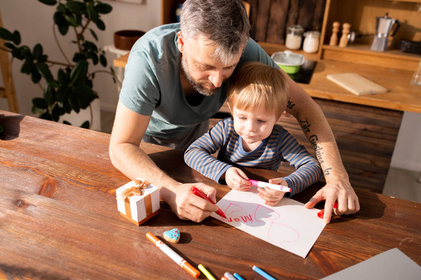 Serious concentrated bearded father and cute son standing at wooden table with markers and gift box and making Valentines card for mother - Фото, зображення