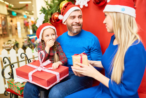 Sorrindo homem e mulher com filhinha em chapéu de Papai Noel dando presentes uns aos outros comemorando o Natal
 - Foto, Imagem