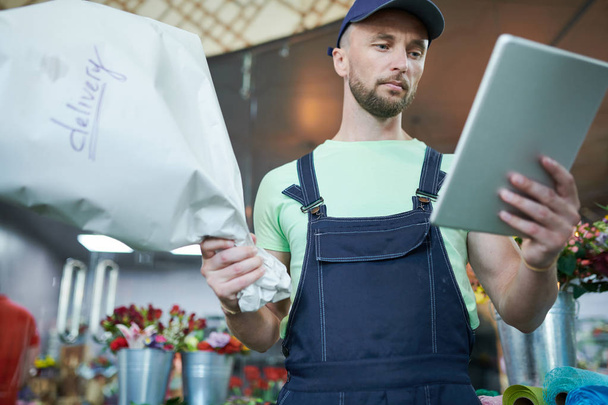 Lage hoek portret van knappe Delivery Guy Holding digitale Tablet en boeket controleren bestelinformatie in bloemenwinkel - Foto, afbeelding