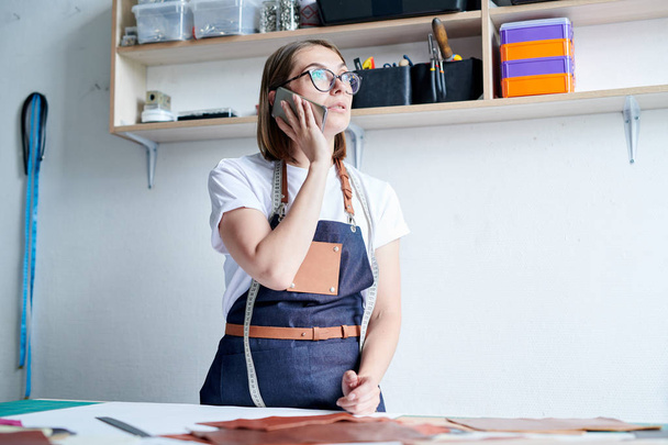 Νεαρή γυναίκα ράφτρα με ποδιά στέκεται στο εργαστήριο και έχει μια επαγγελματική συνομιλία στο τηλέφωνο - Φωτογραφία, εικόνα