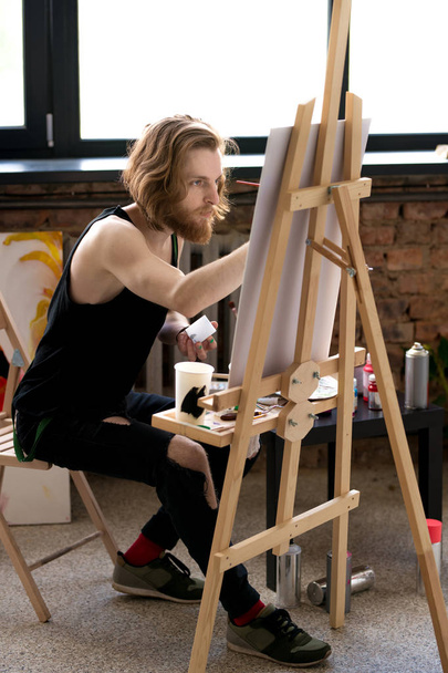 Side view full length portrait of handsome long haired artist  painting on easel in art studio - Photo, Image