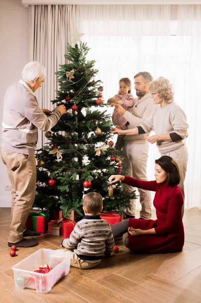 Warm-toned full length portrait of big happy family decorating Christmas tree with two adorable kids and grandparents - Zdjęcie, obraz