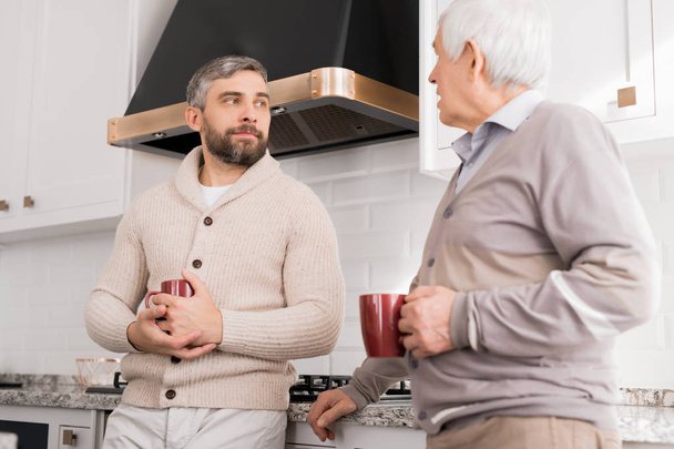 Porträt eines bärtigen erwachsenen Mannes, der zu Hause in der Küche mit seinem Vater spricht - Foto, Bild