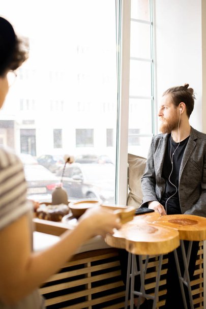 Pensive handsome hipster young bearded man in jacket sitting on window-sill and looking out window in cafe while waitress carrying coffee to him - Photo, Image