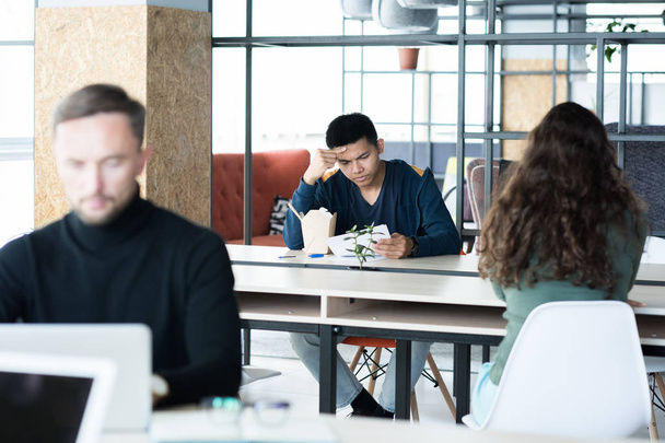 Poważny przemyślany młody Asian Man analizując papiery podczas czytania badań i siedząc przy stole z chińskim jedzeniem, ludzie pracujący i jedzący w otwartej przestrzeni biurowej - Zdjęcie, obraz