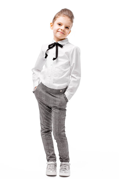 Beautiful little girl in white shirt and gray pants standing near white wall and posing like model - Fotoğraf, Görsel