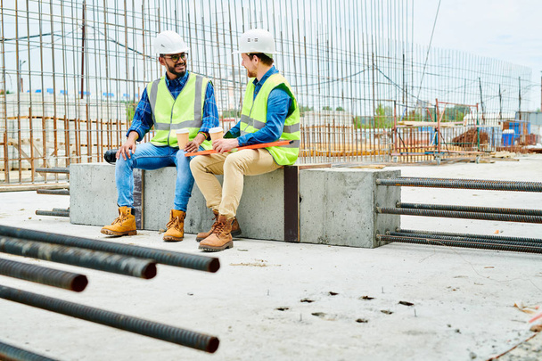 Full length portrait of two workers wearing hardhats  chatting while enjoying coffee break on construction site - Photo, Image