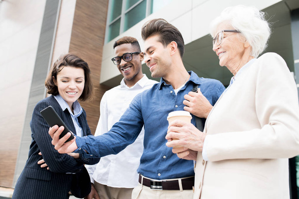 Colegas de negócios multiétnicos confiantes alegres que estão ao ar livre e rindo ao usar o aplicativo móvel conveniente para negócios
 - Foto, Imagem
