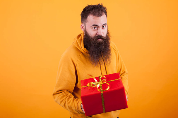 Muotokuva hauska mies pitkä parta osoittaa punainen lahjapaketti keltaisella taustalla
 - Valokuva, kuva