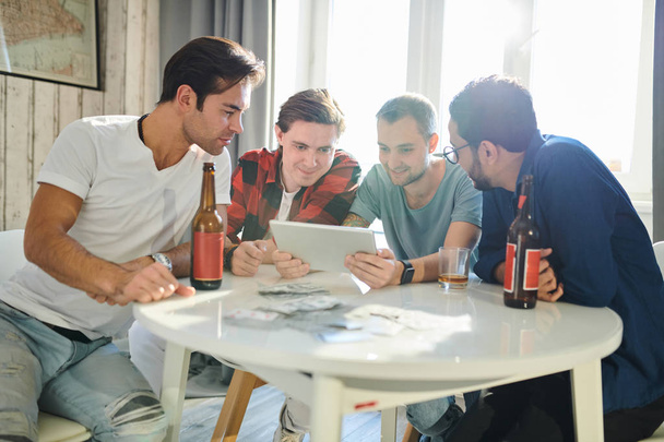 Fiatal férfiak ülnek az asztalnál sört isznak és online kommunikálnak a barátjukkal, vagy digitális táblagéppel online ételrendelést készítenek. - Fotó, kép