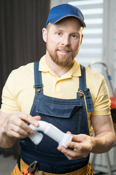 Smiling handsome young bearded plumber in blue cap holding pipe and looking at camera while standing in modern flat - Foto, afbeelding