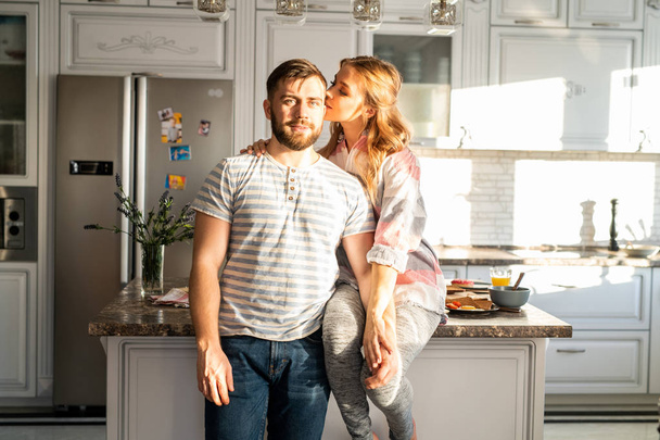Porträt eines modernen glücklichen Paares, das zu Hause posiert, Frau küsst Ehemann, während sie auf dem Küchentisch sitzt - Foto, Bild