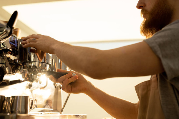 Lähikuva vakava kiireinen parrakas barista mies esiliina liittämällä portafilter espresso koneen keittäessään kahvia kahvilassa
 - Valokuva, kuva
