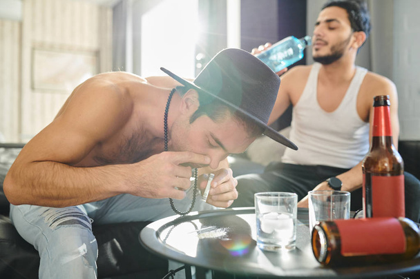 Hombre drogadicto olfateando cocaína con billete enrollado en la fiesta de casa con otro hombre bebiendo vodka en el fondo
 - Foto, imagen