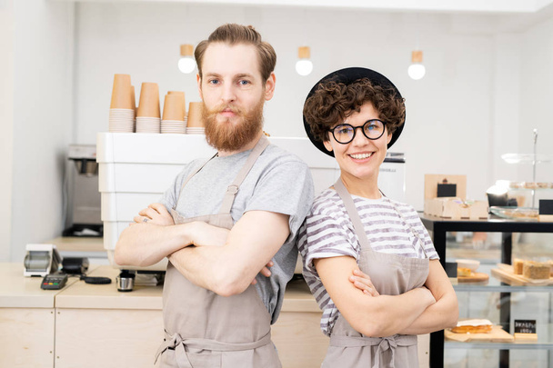 Positive confident hipster couple of baristas in aprons standing against espresso machine and looking at camera - Photo, Image